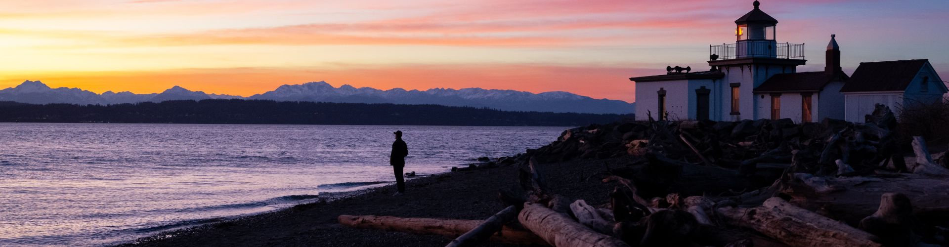 Photo of Discovery Park in Seattle, WA. A man standing on a rocky beach on the Puget Sound in front of a lighthouse with a beautiful multi-colored sunset,